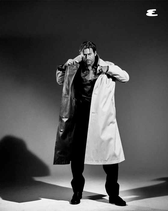 Glen Powell putting on a long coat in a studio portrait with dramatic lighting against a plain backdrop for Esquire U.S.