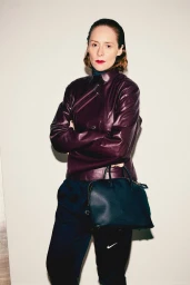 Woman in burgundy leather jacket and black trousers holding a dark handbag against a plain background for Interview Magazine