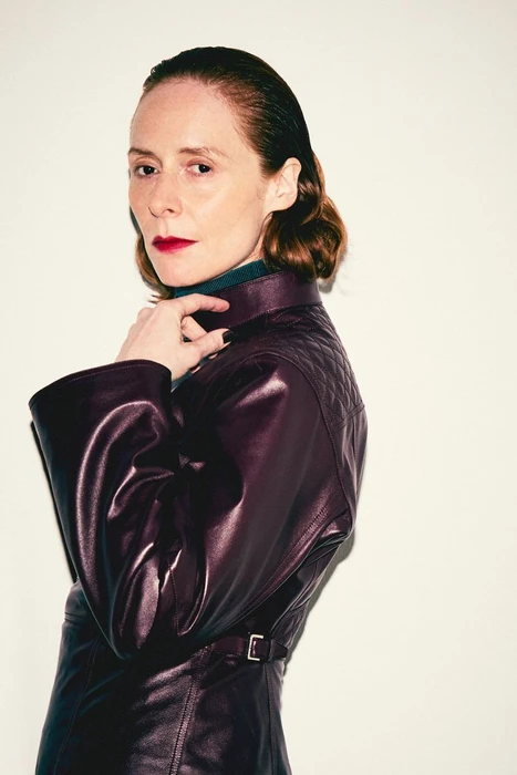 Woman wearing a dark purple leather jacket and dark lipstick against a plain white background