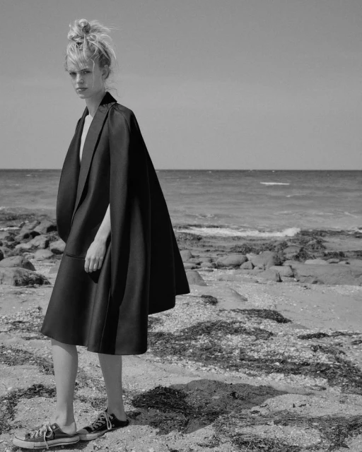 A woman in a dark coat stands on a rocky beach in a black and white fashion shot, with waves and a clear sky in the background.