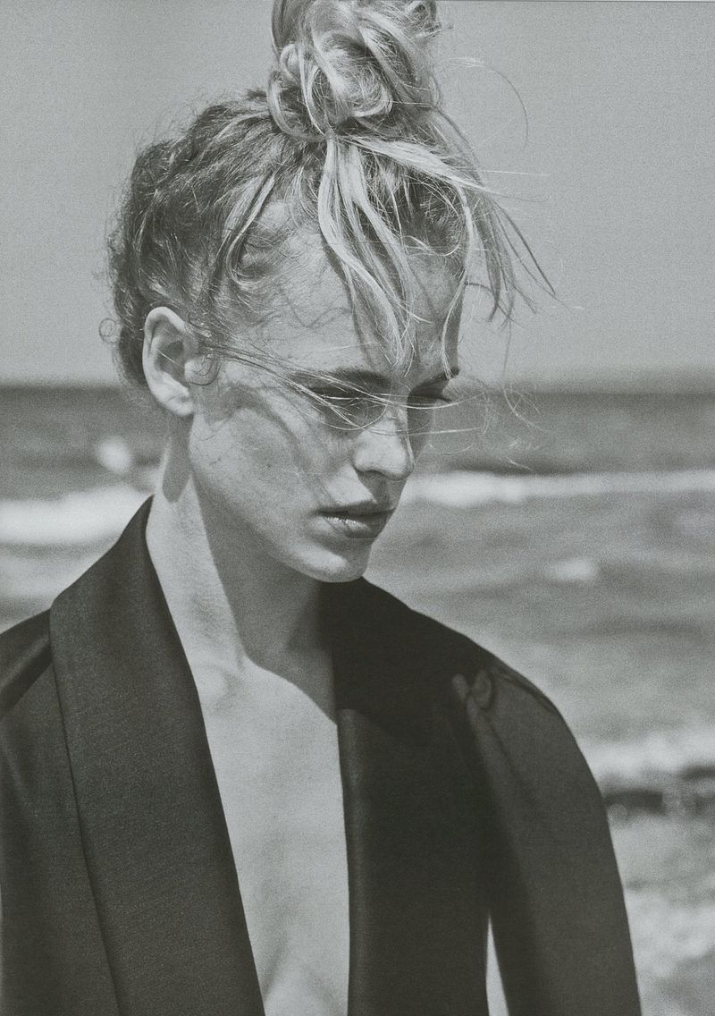 Monochrome portrait of a young woman with a hair bun, wearing a dark jacket, and standing in front of a blurred sea.