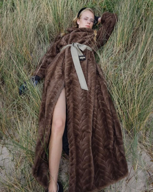 Woman in a long brown faux fur coat lying in tall grass, wearing eyeglasses and a black headband with a light green belt.