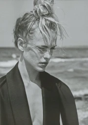 Black and white portrait of a woman with windblown hair tied in a bun, wearing a tailored dark jacket against an ocean horizon.