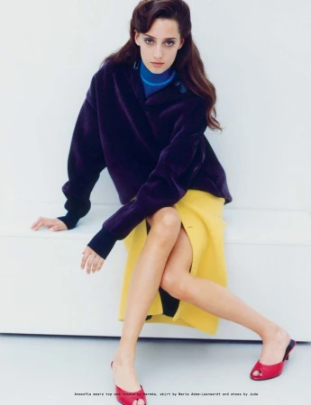 Model Anasofia Negrutsa seated wearing a purple jacket, blue turtleneck, yellow skirt and red mules on a white seamless backdrop