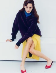 Model Anasofia Negrutsa seated wearing a purple jacket, blue turtleneck, yellow skirt and red mules on a white seamless backdrop
