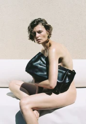 Model Bibi Breslin holding a black leather handbag while seated against a textured wall for Purple Magazine.