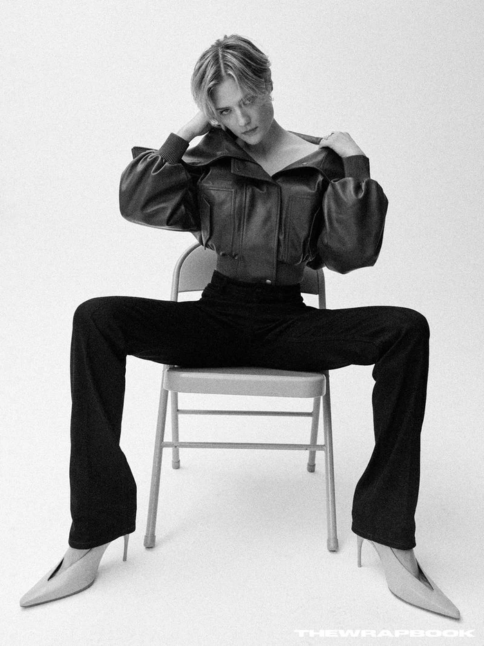 Louisa Jacobson seated on a folding chair wearing a dark leather jacket and dark trousers against a white seamless backdrop.