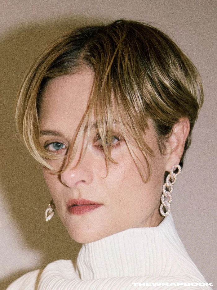 Close-up portrait of Louisa Jacobson wearing a white ribbed turtleneck and diamond link earrings against a plain background