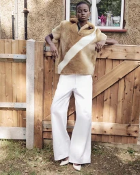 Model leaning against a wooden fence wearing a fluffy brown top and white trousers