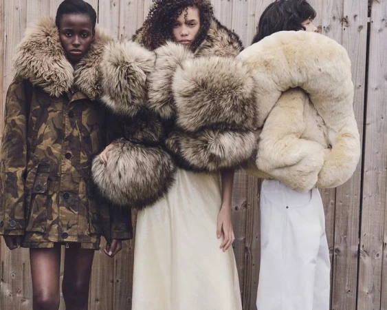 Three models wearing oversized fur-trimmed outerwear standing against a wooden slat wall for W Magazine editorial.