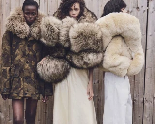 Three models wearing oversized fur-trimmed outerwear standing against a wooden slat wall for W Magazine editorial.