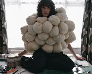 Model wearing a voluminous padded white sculptural garment seated at a cutting table in a workroom.