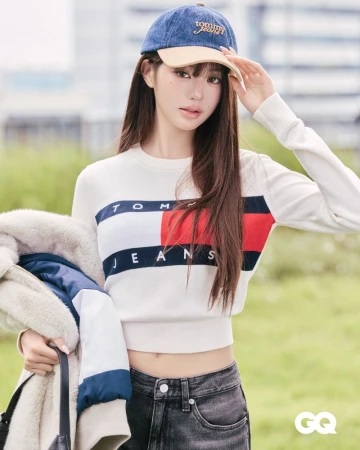 Young woman wearing a denim baseball cap, white crewneck sweater, and dark jeans holding a winter jacket outdoors.