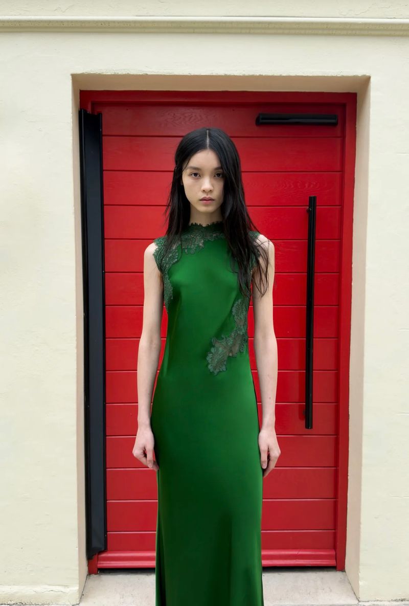 Model in a green lace-trimmed sleeveless dress standing in front of a bright red slatted door for Self-Portrait Spring 2027.