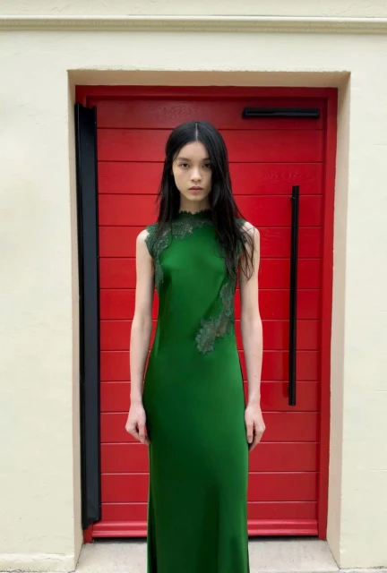 Model in a green lace-trimmed sleeveless dress standing in front of a bright red slatted door for Self-Portrait Spring 2027.