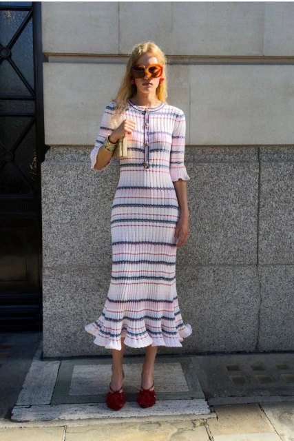 Model wearing a striped knit dress and tinted sunglasses standing in front of a stone building exterior.