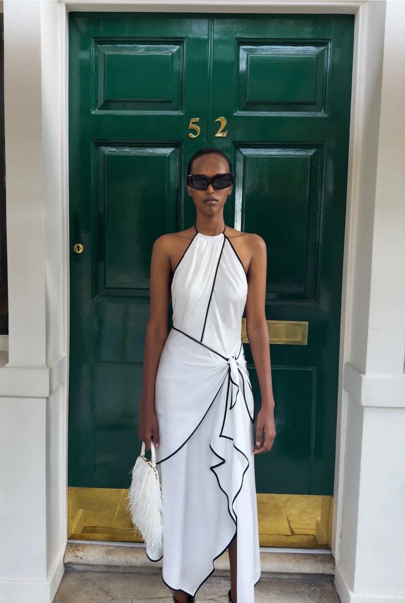 Model standing in a white halter dress with black trim in front of a green double door featuring number 52