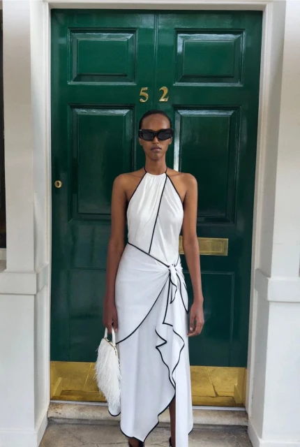Model standing in a white halter dress with black trim in front of a green double door featuring number 52