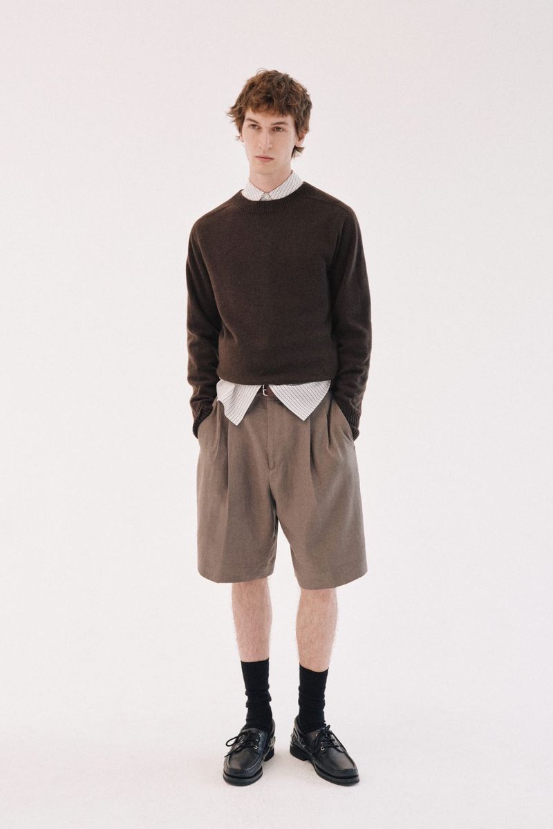 Model wearing a dark brown sweater over a striped shirt and tan shorts with black socks and shoes against a white backdrop.