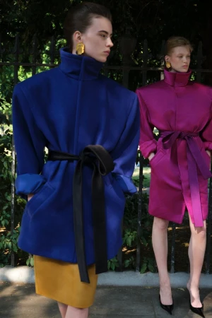 Two women model blue and magenta coats with belts and gold earrings against a green leafy background. Fashion shot, eye-level angle.