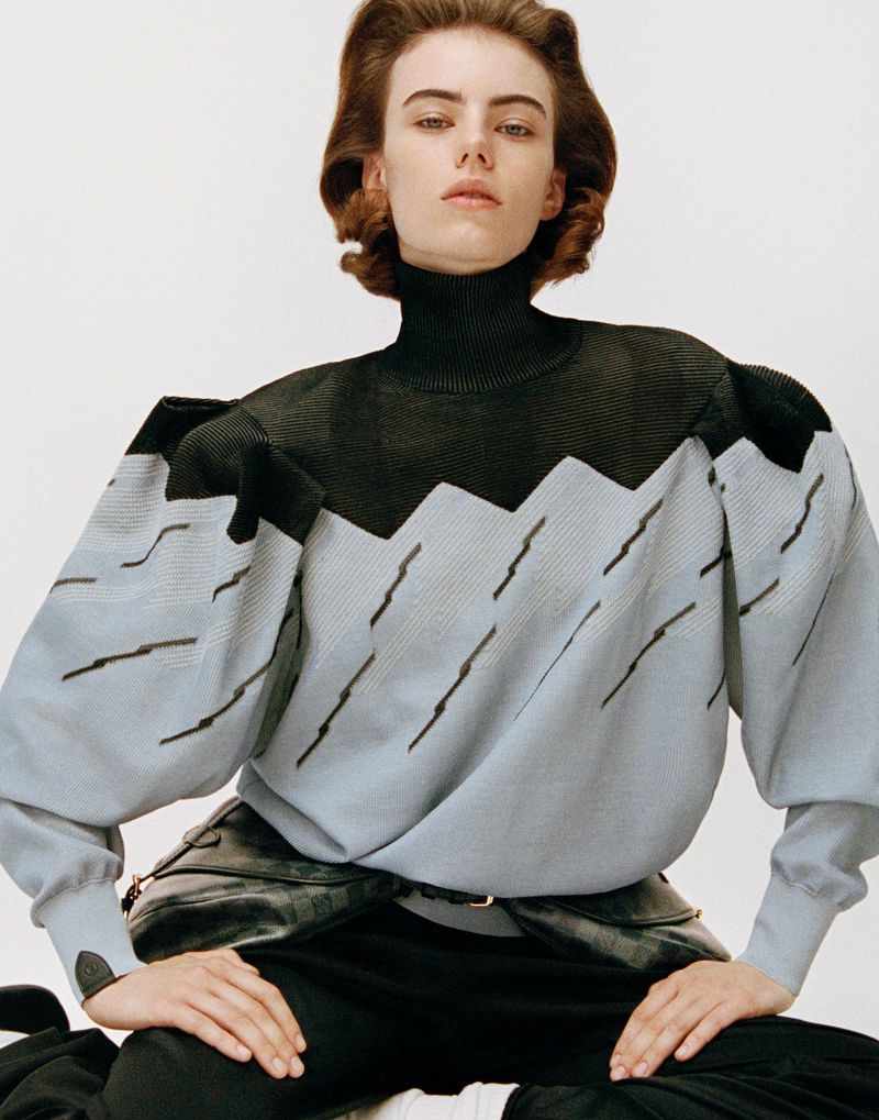 A young woman poses seated indoors with a neutral expression. She is wearing a black and light blue patterned sweater and dark pants.