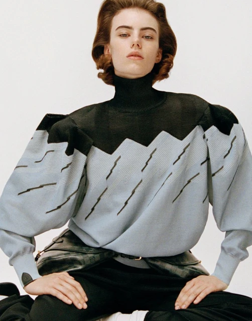 A young woman poses seated indoors with a neutral expression. She is wearing a black and light blue patterned sweater and dark pants.