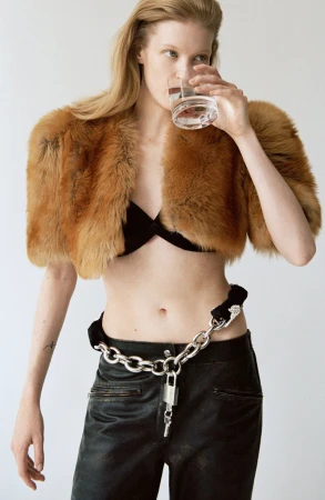 A blonde woman in a fur shrug drinks water, wearing black leather pants and a bra top in a studio setting with a light background.