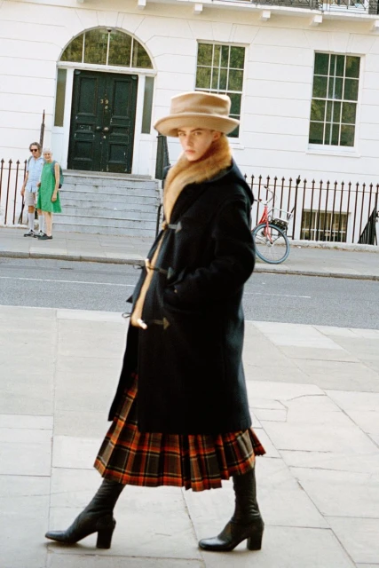 A fashionable woman in a black coat, plaid skirt, and hat walks down a city sidewalk near a white building on a sunny day.