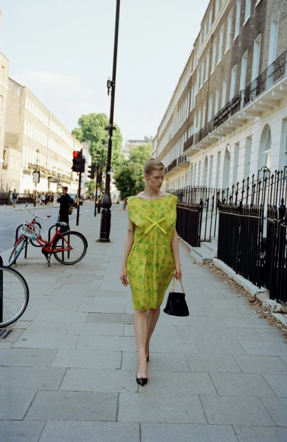 Woman in yellow dress walks on city sidewalk with a black purse, brick buildings, trees and traffic lights in the background on a sunny day.