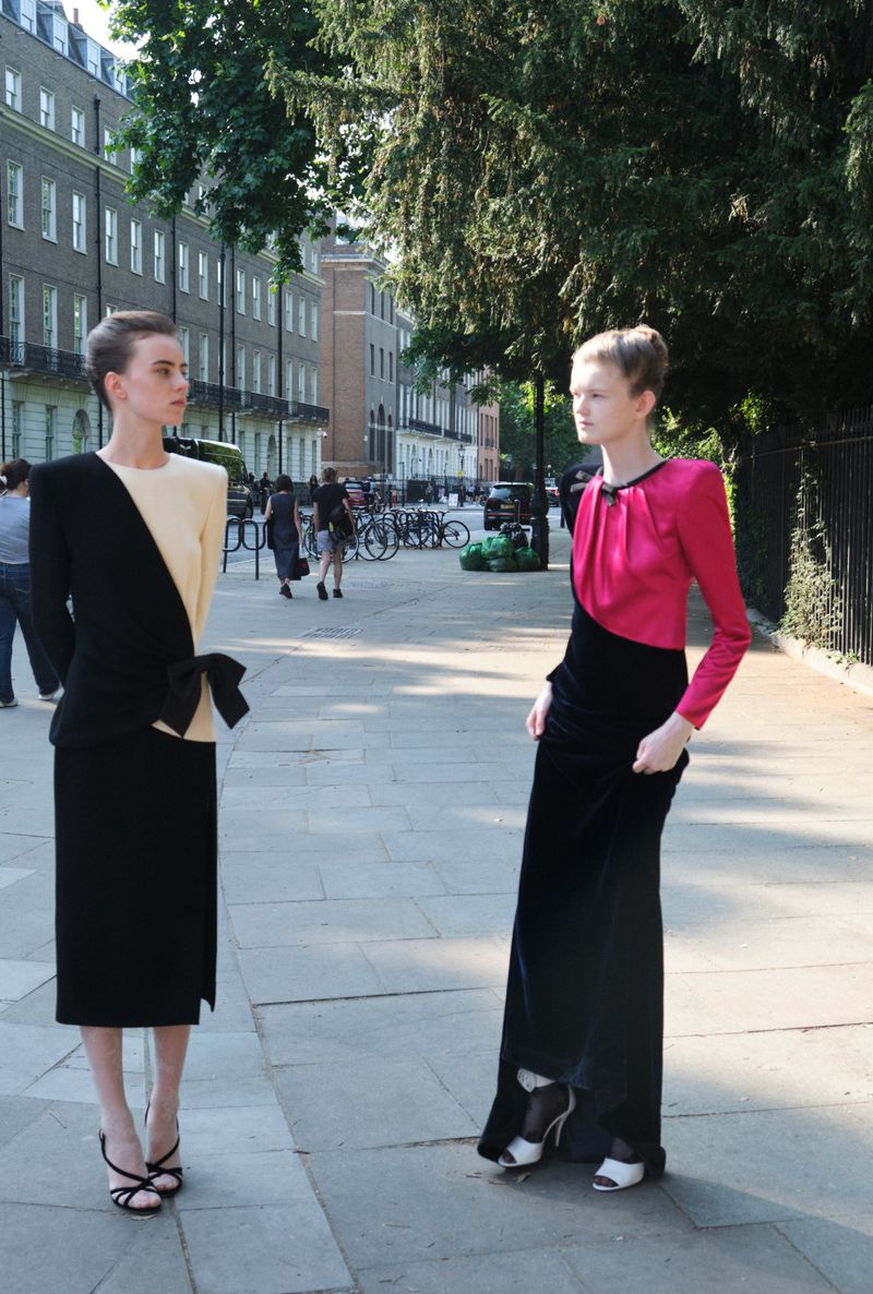 Two fashion models pose outdoors on a city sidewalk wearing elegant, vintage-inspired color-blocked dresses during daylight hours.