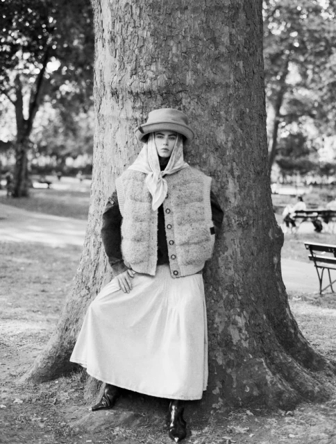 Black and white outdoor portrait of a young woman in vintage clothing, wearing a hat and scarf, leaning against a large tree.