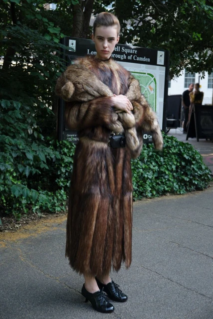Woman in full-length fur coat stands on paved path in front of green foliage and signage in an outdoor setting, looking at the camera.
