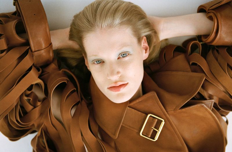 Woman with fair skin poses in a brown leather trench coat with a gold buckle, laying against a white background in a studio setting.