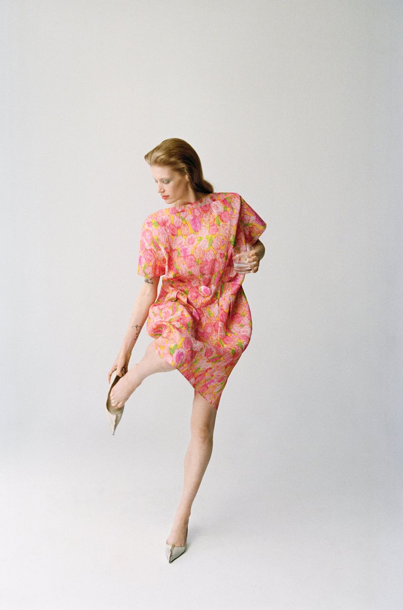 A woman in a pink floral dress and silver heels stands against a white background, her back turned toward the viewer.
