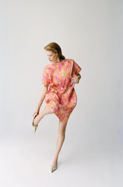 A woman in a pink floral dress and silver heels stands against a white background, her back turned toward the viewer.