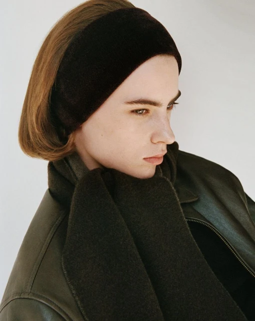 Portrait of a young adult woman in a dark brown headband and scarf, wearing a dark leather jacket against an off-white background.