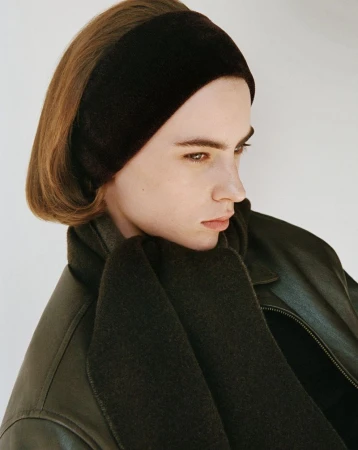 Portrait of a young adult woman in a dark brown headband and scarf, wearing a dark leather jacket against an off-white background.