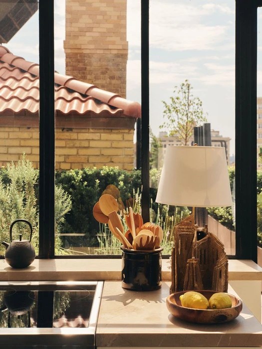 Kitchen counter with a bowl of lemons, wooden utensils, lamp, and black teapot in front of a large window view.