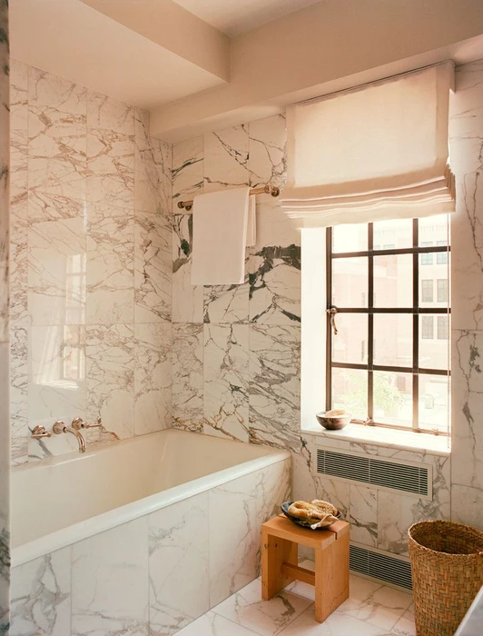 Luxurious marble bathroom featuring a bathtub and window with roman shades photographed for Architectural Digest.