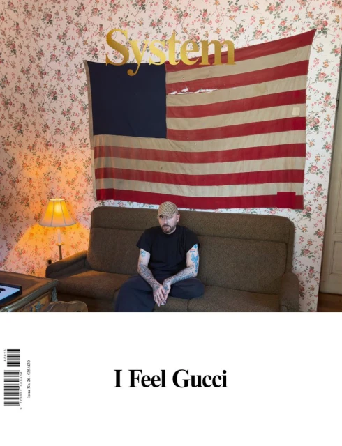 Demna seated on a brown sofa beneath an American flag in a floral wallpaper room for System Magazine 26 cover photographed by Juergen Teller.