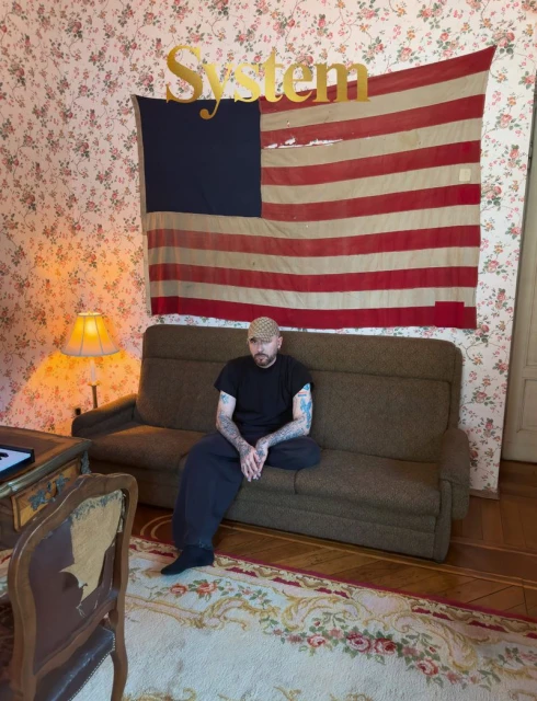 Man with tattoos seated on a brown sofa beneath an American flag and floral wallpaper for System Magazine