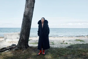 Model standing outdoors near a tree on a coastal beach wearing a long black coat and red boots