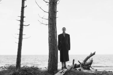 Model standing between trees near a body of water wearing a long dark coat and statement necklace