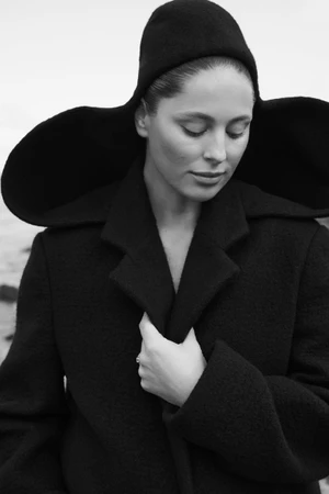Black and white portrait of a woman wearing a dark wide-brimmed hat and coat, holding her lapel outdoors.