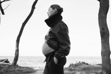 Pregnant woman in leather jacket standing outdoors near trees and water