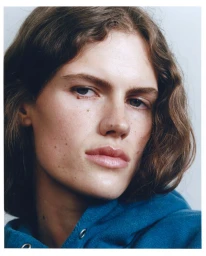 Close-up portrait of a person with wavy brown hair wearing a blue hoodie against a plain white wall