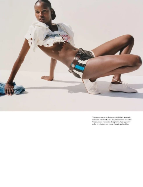 Fashion shot of Black female model in a pose on a white surface, wearing a white crop top, print shorts, and off-white shoes.