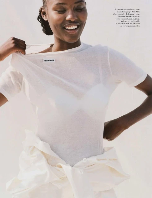 Smiling Black woman wearing a sheer white top and baggy pants in a studio, looking cheerful and fashionable in a close-up shot.