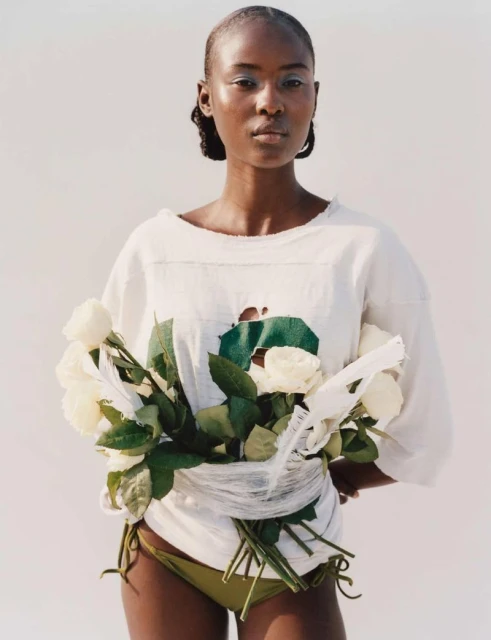 Striking portrait of a Black woman in a white top and bikini bottom, holding white roses against a clean white backdrop.