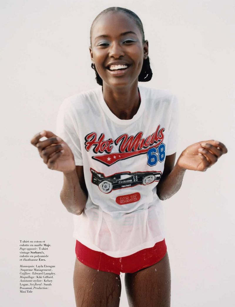 A smiling African-American woman in a wet t-shirt with a racecar graphic and red shorts poses in front of a white background.